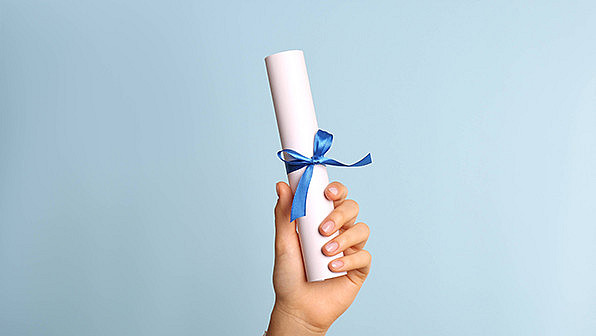 Eine Hand hält vor hellblauem Hintergrund ein zusammengerolltes Zertifikat hoch, das mit einer blauen Schleife zusammengebunden ist.