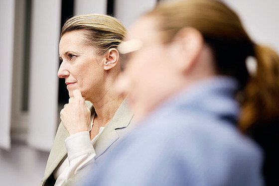 Eine Frau mit zurückgebundenem blonden Haar sitzt im Profil, stützt ihr Kinn auf die Hand und wirkt nachdenklich. Eine weitere Person mit braunem Haar zu einem Pferdeschwanz ist im Vordergrund unscharf zu sehen.