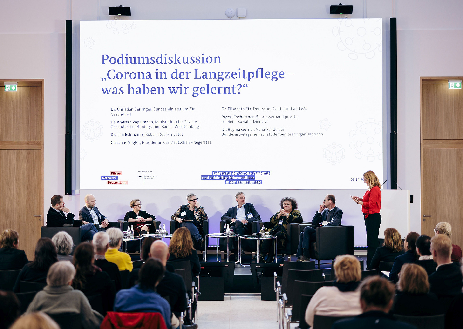Eine Podiumsdiskussion in einem Konferenzraum mit neun Personen auf der Bühne. Auf einer großen Leinwand wird das Thema auf Deutsch über Langzeitpflege und Lehren aus der Pandemie gezeigt. Im Vordergrund sitzen die Zuschauer, eine Frau steht auf und spricht.