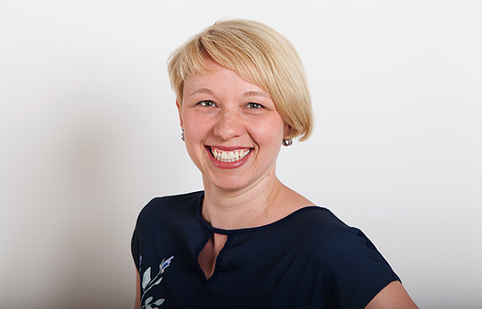 Eine lächelnde Person mit kurzen blonden Haaren trägt ein marineblaues Oberteil mit einem kleinen Blumenmuster auf dem Ärmel und steht vor einem schlichten weißen Hintergrund.