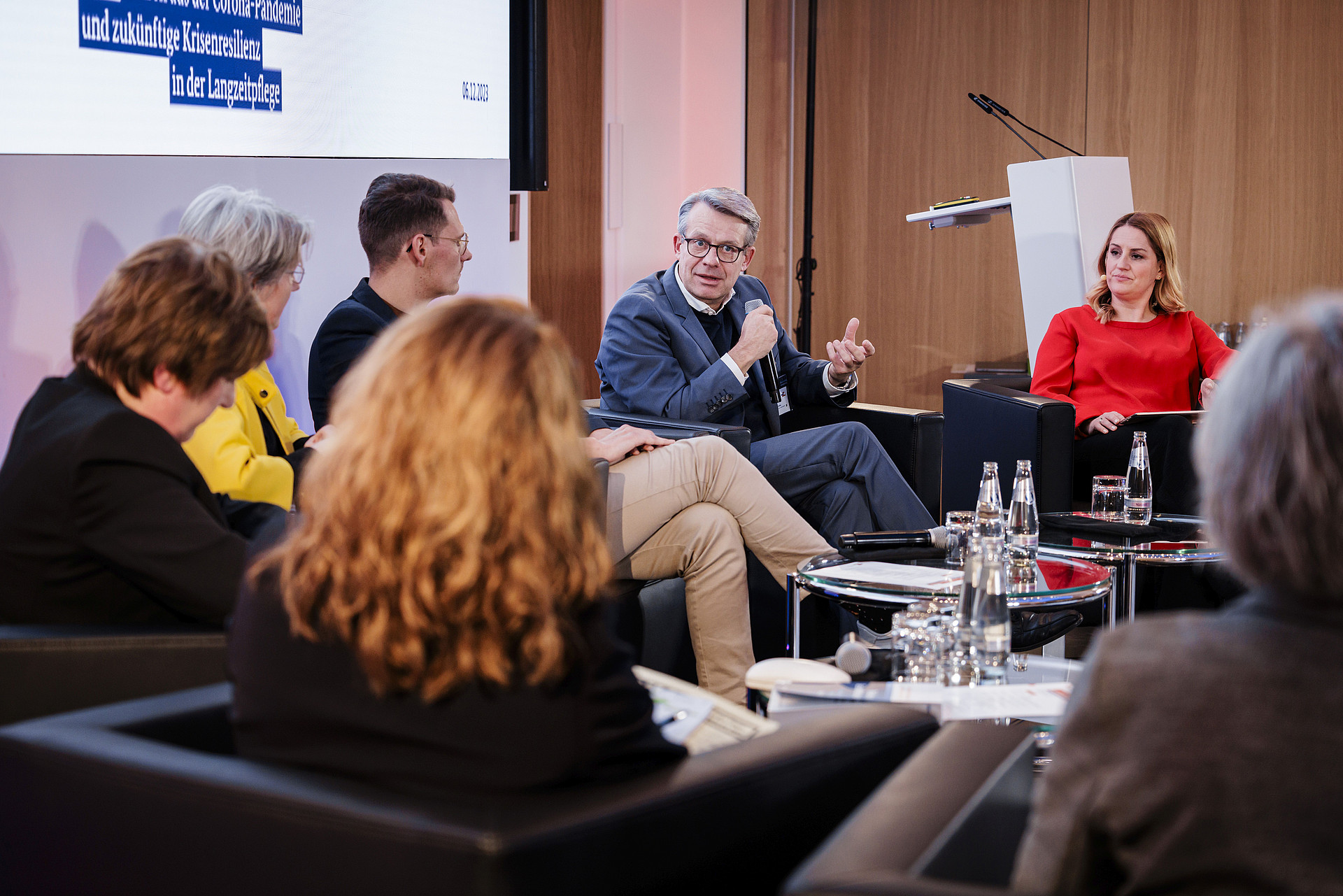 Eine Gruppe von Menschen sitzt im Halbkreis und nimmt an einer Podiumsdiskussion teil. Ein Mann hält ein Mikrofon, während die anderen aufmerksam zuhören. Eine Frau in einem roten Oberteil sitzt neben ihm. Das Ambiente wirkt professionell, mit Tischen, Wasserflaschen und einem Rednerpult.