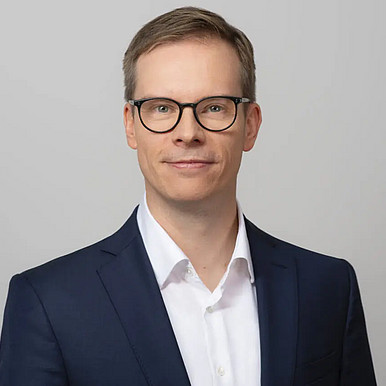 Ein Mann mit Brille und marineblauem Anzug mit weißem Hemd steht vor einem schlichten hellgrauen Hintergrund und blickt mit einem leichten Lächeln in die Kamera.