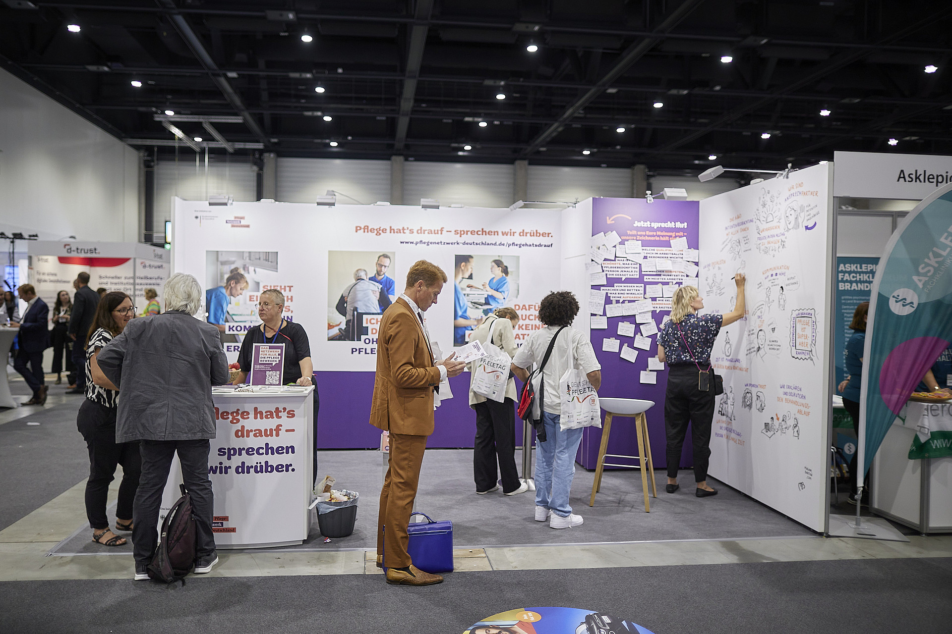 Menschen auf einer Tagung interagieren mit Exponaten; manche lesen die Displays, während andere auf ein großes Whiteboard schreiben. Im Hintergrund sind Standaufbauten und Beschilderungen in einer geräumigen Halle mit Deckenbeleuchtung zu sehen.