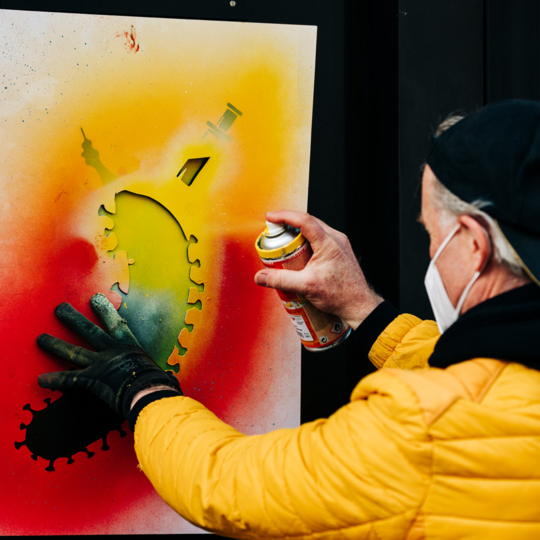 Eine Person in einer gelben Jacke und einer schwarzen Kappe verwendet Sprühfarbe auf einer Schablone, die die Form eines Virus mit einer Spritze darstellt. Die Farben sind leuchtend, darunter Rot, Gelb und Grün. Die Person trägt Handschuhe und eine Gesichtsmaske und konzentriert sich auf das Kunstwerk.
