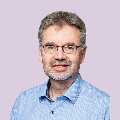 Ein Mann mittleren Alters mit kurzen grauen Haaren, Brille und gestutztem Bart, der ein hellblaues Hemd mit Kragen trägt, lächelt vor einem schlichten hellvioletten Hintergrund in die Kamera.