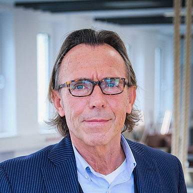 Ein Mann mit Brille und schulterlangem Haar trägt einen blau gestreiften Anzug und ein hellblaues Hemd. Er steht in einem hellen Büro mit großen Fenstern im Hintergrund.