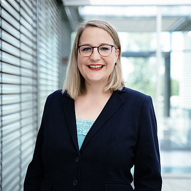 Eine Person mit schulterlangen blonden Haaren und Brille lächelt und trägt einen dunklen Blazer über einem hellblauen Oberteil. Sie steht in einer modernen Inneneinrichtung mit Metall- und Glaselementen.