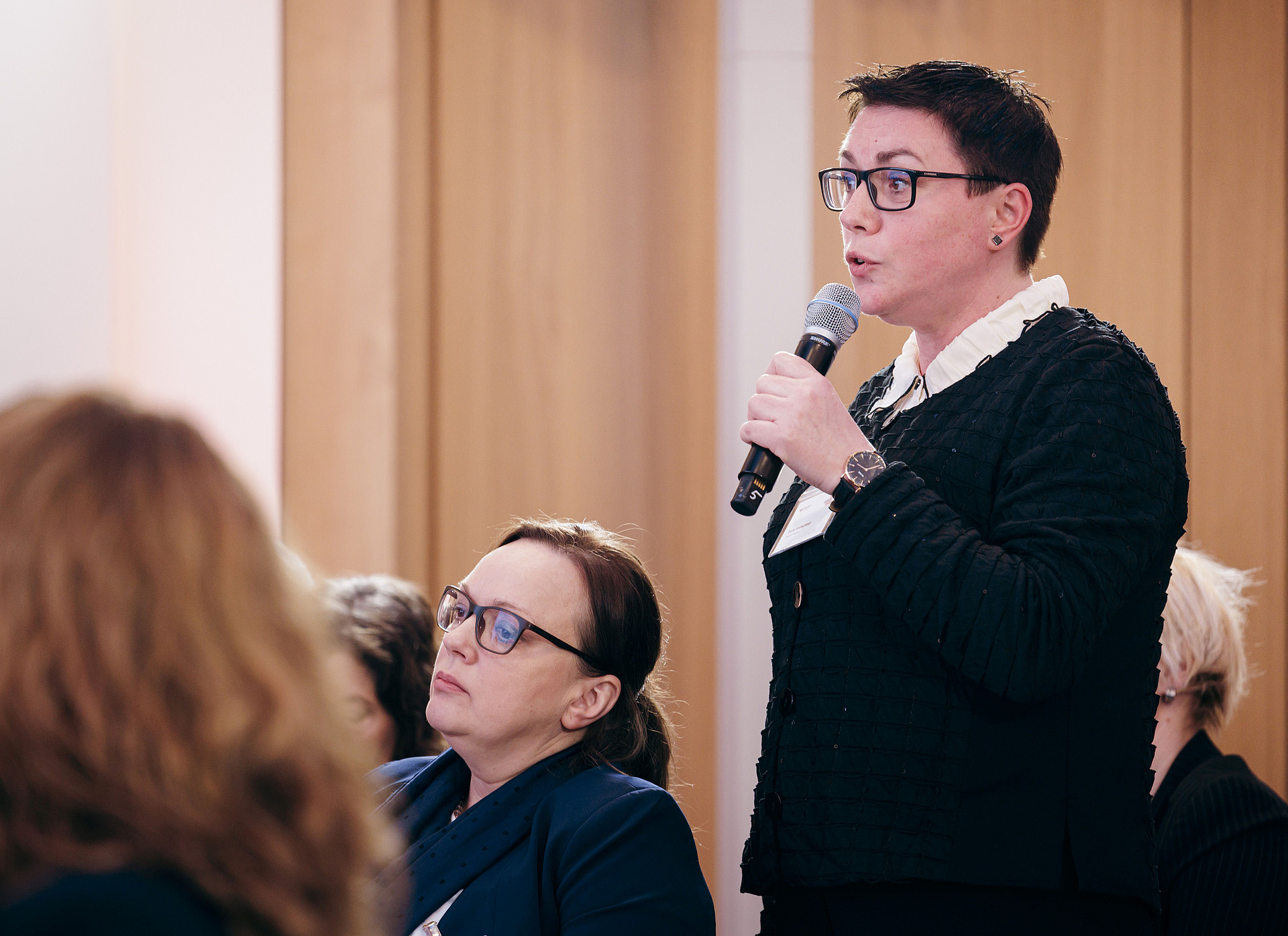 Eine Person steht mit einem Mikrofon da und spricht während einer Veranstaltung. Eine andere Person sitzt im Vordergrund und hört aufmerksam zu. Beide sind formelle Kleidung und die Szene scheint sich in einem Innenbereich mit Holzvertäfelung im Hintergrund zu befinden.