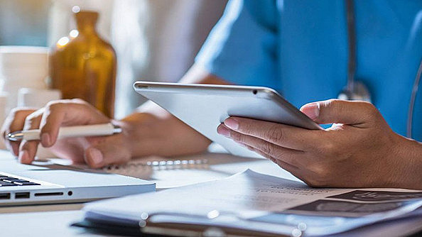 Eine Person im OP-Kittel benutzt ein Tablet und schreibt auf Papier an einem Schreibtisch. In der Nähe liegen ein Laptop, ein Stift und Dokumente. Sonnenlicht erhellt den Arbeitsbereich und suggeriert eine professionelle Umgebung.