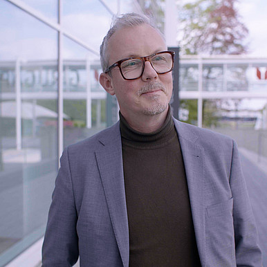 Ein Herr mit grauem kurzem Haar und Brille ist vor einen Glasgebäude zu sehen. Er schaut zur Kamera. Es ist Prof. Isfort.