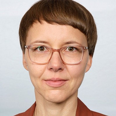 Eine Person mit kurzen braunen Haaren, einer großen, klaren Brille und einem braunen Oberteil blickt vor einem hellen, schlichten Hintergrund direkt in die Kamera.