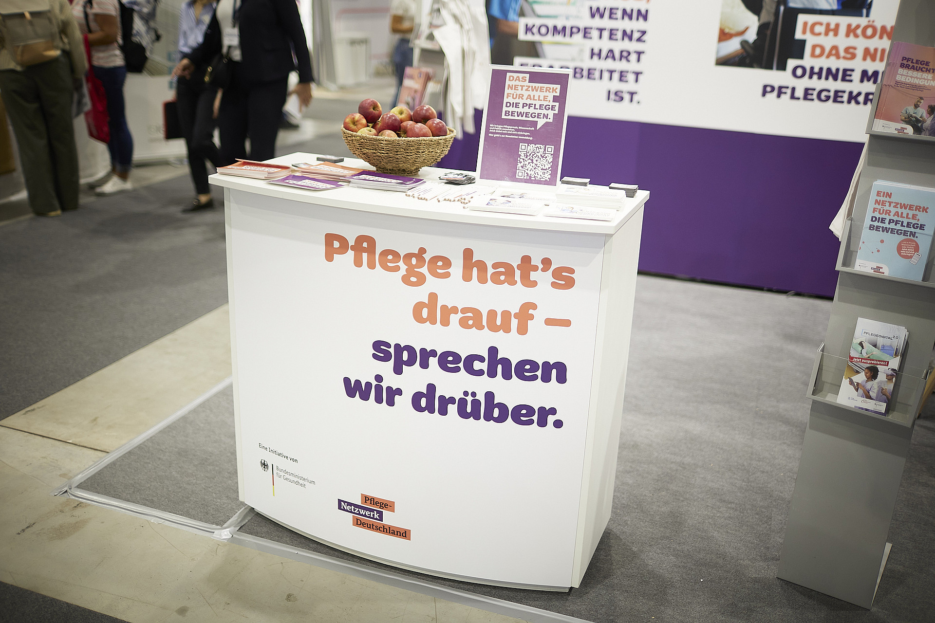 An einem Informationsstand auf einer Tagung werden Broschüren und ein Korb mit Äpfeln ausgestellt. Der Text auf dem Stand lautet: „Pflege hat’s drauf – sprechen wir drüber“, was darauf schließen lässt, dass der Schwerpunkt auf Gesundheit und Krankenpflege liegt.