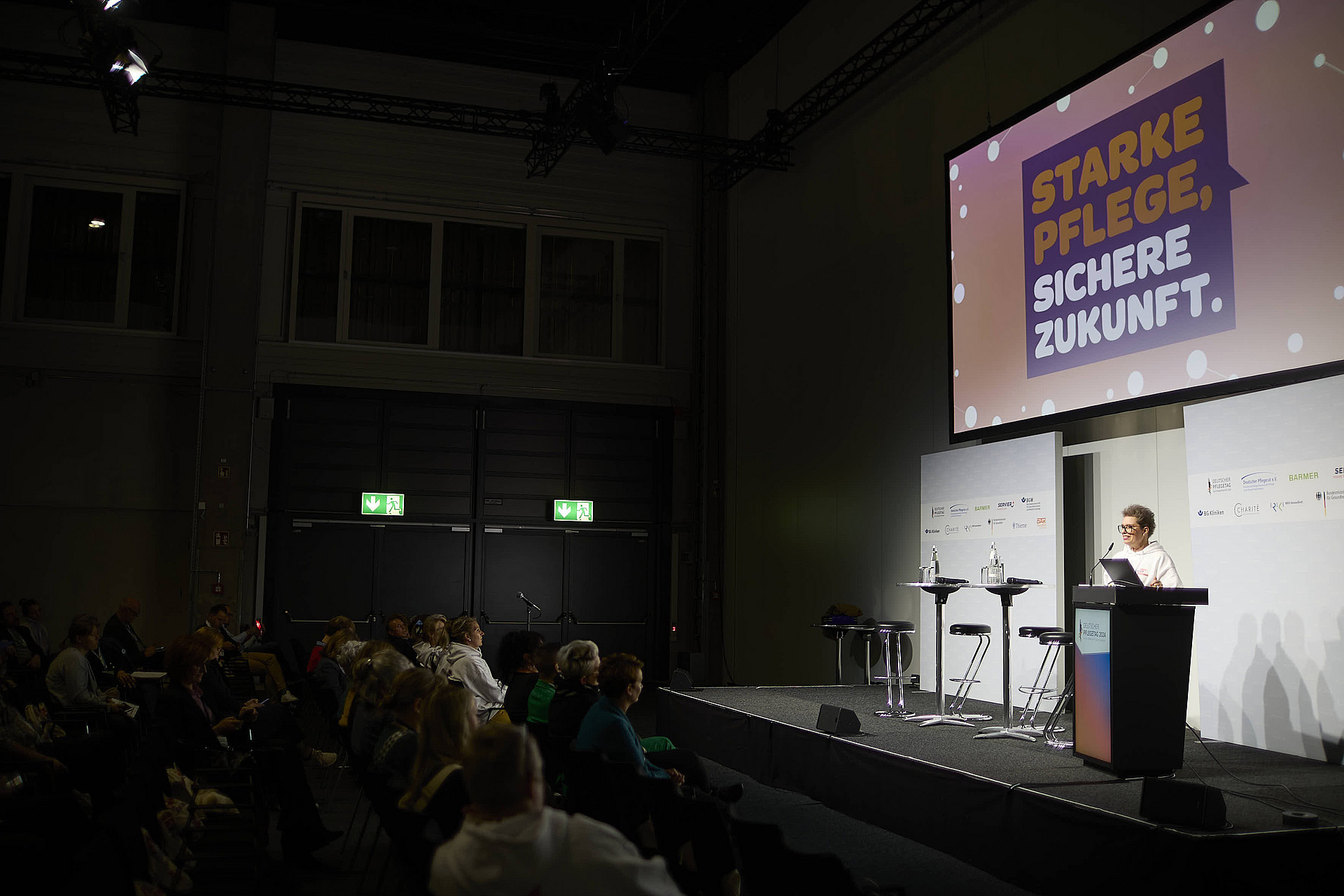 Frau steht an einem Rednerpult bei einer Veranstaltung. Davor sitzt Publikum. An der Wand ist groß "Starke Pflege sichere Zukunft" auf einem Bildschirm zu lesen.