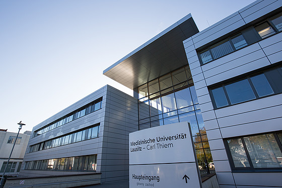 Modernes Gebäude mit Glaseingang und Metallfassade; ein Schild davor mit der Aufschrift „Medizinische Universität Lausitz – Carl Thiem“ und „Haupteingang“ weist darauf hin, dass es sich um den Haupteingang einer Medizinischen Universität handelt.