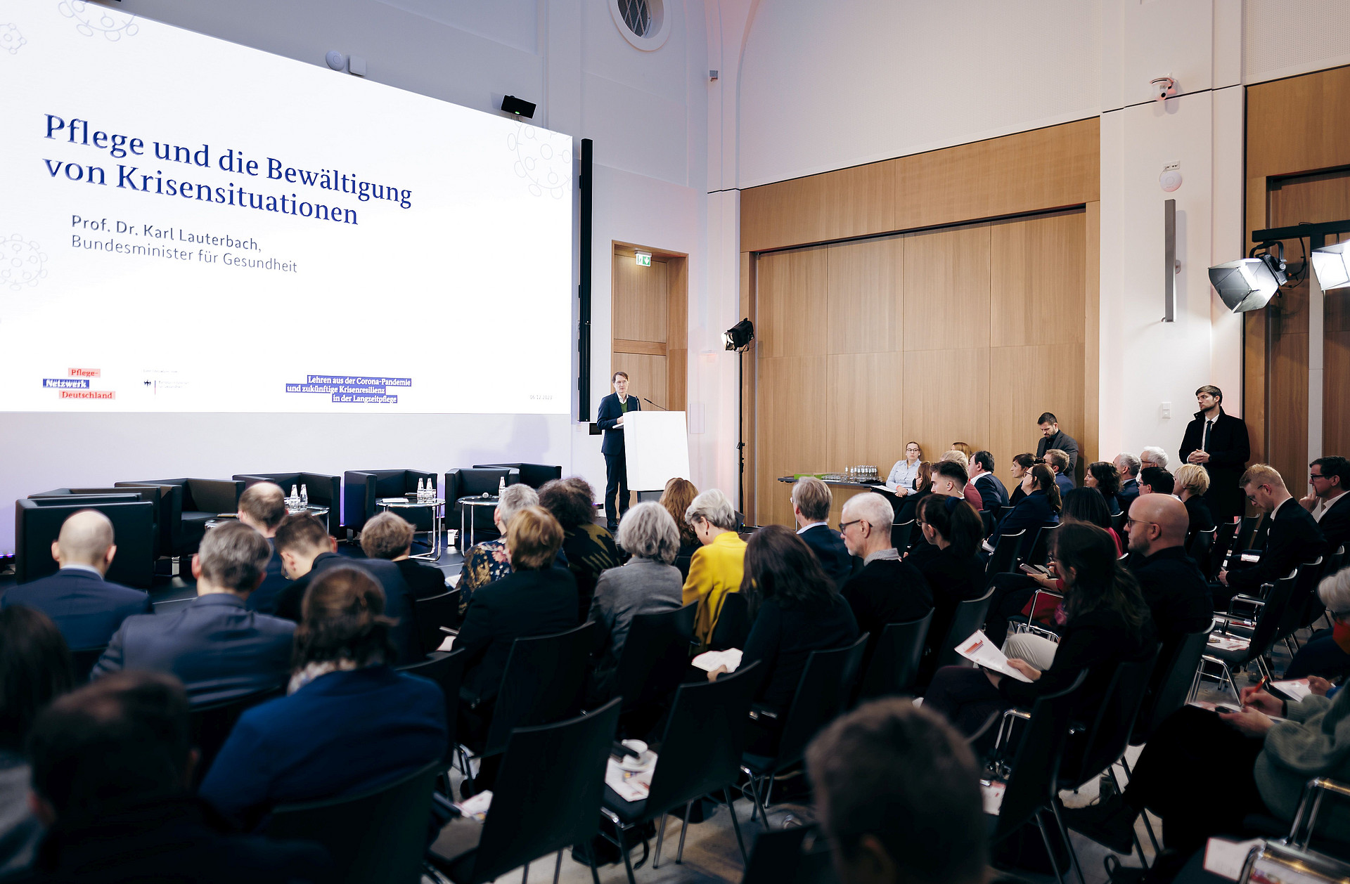 Ein Redner spricht zu einem Publikum in einem Konferenzraum. Auf einem großen Bildschirm läuft ein deutscher Text über Krisenmanagement im Gesundheitswesen. Der Raum ist gut beleuchtet, die Teilnehmer sitzen, einige machen sich Notizen.