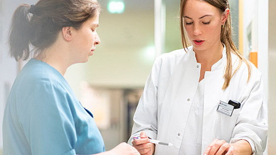 Zwei medizinische Fachkräfte besprechen in einem Krankenhausflur Notizen auf einem Klemmbrett. Einer trägt eine blaue Uniform, der andere einen weißen Kittel und hält einen Stift in der Hand. Die Szene ist hell beleuchtet und der Hintergrund ist unscharf.