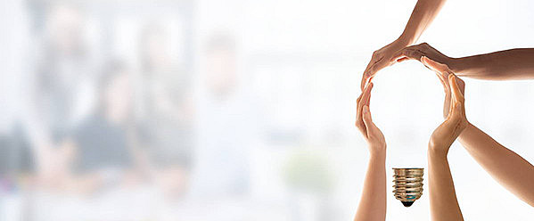 Hände bilden die Form einer Glühbirne mit einem metallischen Glühbirnensockel an der Unterseite. Der Hintergrund ist verschwommen und zeigt möglicherweise Personen oder eine Gruppensituation.