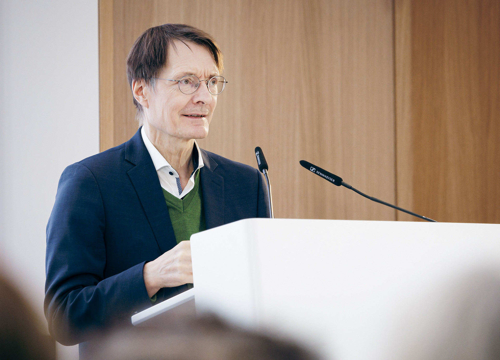 Ein Mann mit Brille und Anzug steht an einem Rednerpult mit Mikrofon und spricht. Es ist Prof. Karl Lauterbach. Er befindet sich in einem Gebäude vor einer Holzwand im Hintergrund.