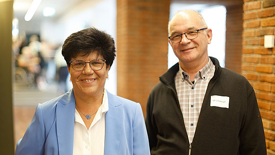 Zwei lächelnde Menschen stehen drinnen. Die Frau links trägt einen hellblauen Blazer und ein weißes Hemd, hat kurze dunkle Haare und eine Brille. Der Mann rechts trägt ein kariertes Hemd und eine dunkle Jacke und hat kurze graue Haare. Im Hintergrund eine Backsteinmauer.