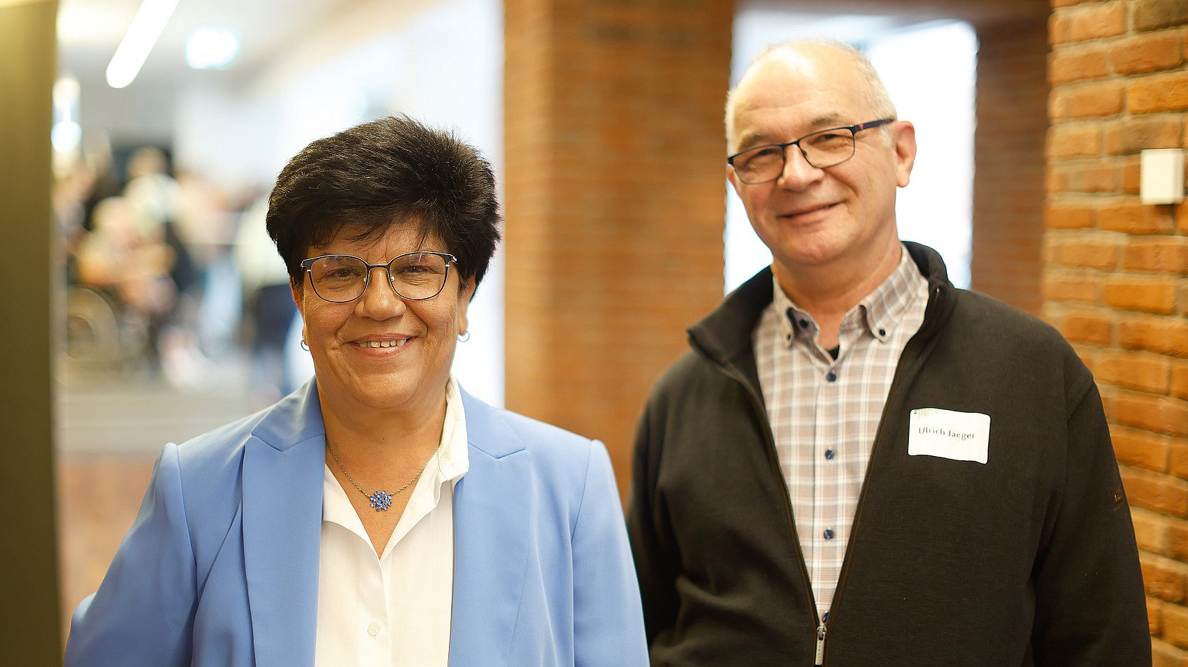 Zwei lächelnde Menschen stehen drinnen. Die Frau links trägt einen hellblauen Blazer und ein weißes Hemd, hat kurze dunkle Haare und eine Brille. Der Mann rechts trägt ein kariertes Hemd und eine dunkle Jacke und hat kurze graue Haare. Im Hintergrund eine Backsteinmauer.