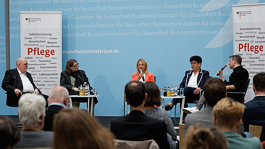 Ein fünfköpfiges Gremium sitzt auf der Bühne und diskutiert. Sie befinden sich in einem Konferenzraum mit einem Publikum vorn und auf beiden Seiten Bannern zum Thema Gesundheitsfürsorge. Im Hintergrund ist ein Text über das deutsche Gesundheitsministerium zu sehen.