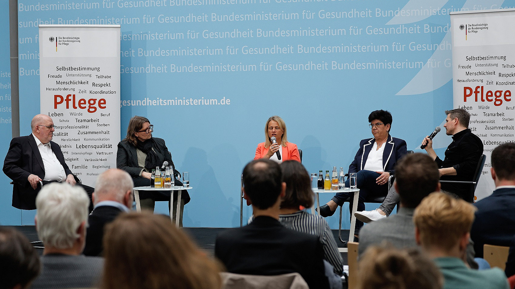 Ein fünfköpfiges Gremium sitzt auf der Bühne und diskutiert. Sie befinden sich in einem Konferenzraum mit einem Publikum vorn und auf beiden Seiten Bannern zum Thema Gesundheitsfürsorge. Im Hintergrund ist ein Text über das deutsche Gesundheitsministerium zu sehen.