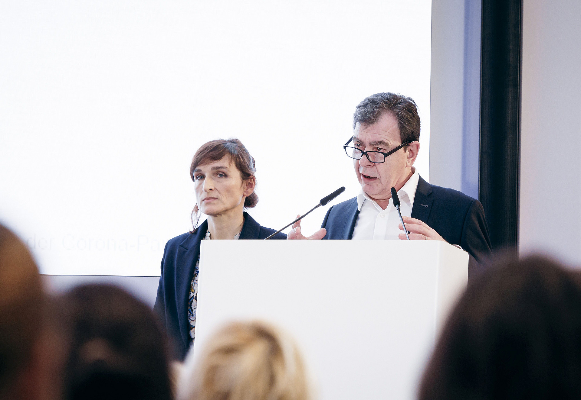 Holger Liljeberg und Dolores Domke stehen an einem Podium und halten eine Präsentation. Der Mann spricht in ein Mikrofon und gestikuliert mit den Händen, während die Frau aufmerksam zuhört. Sie sind in formelle Kleidung gekleidet und im Vordergrund ist ein Publikum zu sehen.