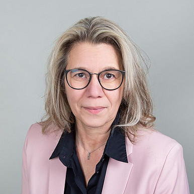 Eine Frau mit schulterlangem blondem Haar, Brille, hellrosa Blazer und schwarzem Hemd posiert vor einem schlichten grauen Hintergrund.