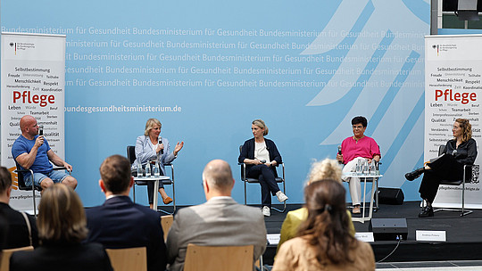 Ein fünfköpfiges Gremium sitzt mit Mikrofonen auf einer Bühne und diskutiert. Im Hintergrund ist ein Text zu sehen, der unter anderem „Bundesministerium für Gesundheit“ enthält. Davor sitzt ein Publikum und hört zu. Zwei Banner mit dem Wort „Pflege“ sind zu sehen.