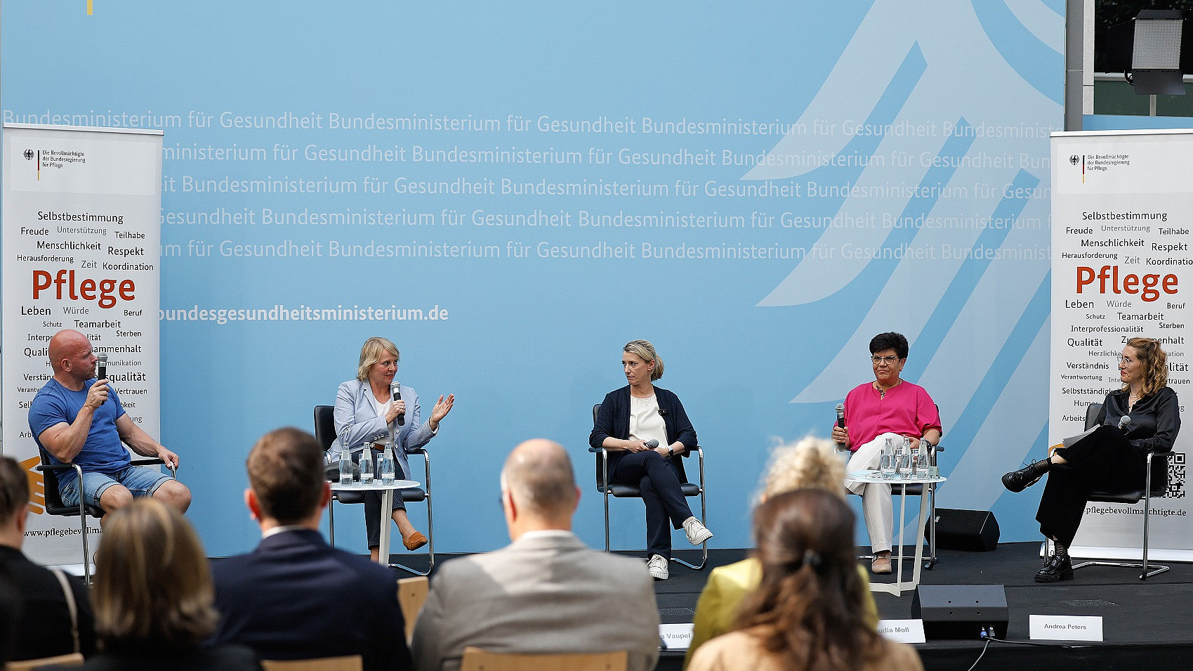 Ein fünfköpfiges Gremium sitzt mit Mikrofonen auf einer Bühne und diskutiert. Im Hintergrund ist ein Text zu sehen, der unter anderem „Bundesministerium für Gesundheit“ enthält. Davor sitzt ein Publikum und hört zu. Zwei Banner mit dem Wort „Pflege“ sind zu sehen.