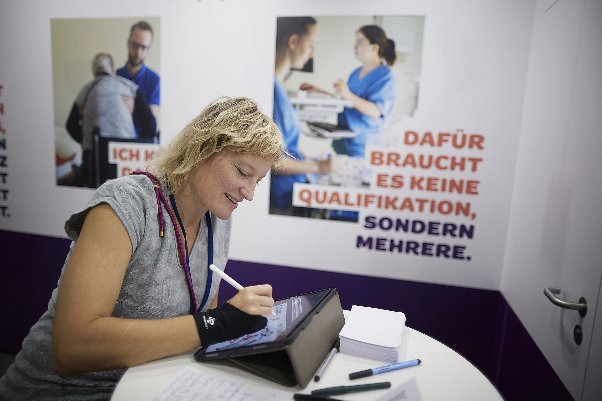 Eine Frau sitzt an einem Tisch und schreibt auf einem Tablet. Neben ihr liegen Notizblöcke und Papiere. Sie trägt eine Handgelenkstütze. Hinter ihr sind Plakate mit deutschen Texten zu sehen, auf denen Menschen in Gesundheitseinrichtungen zu sehen sind.