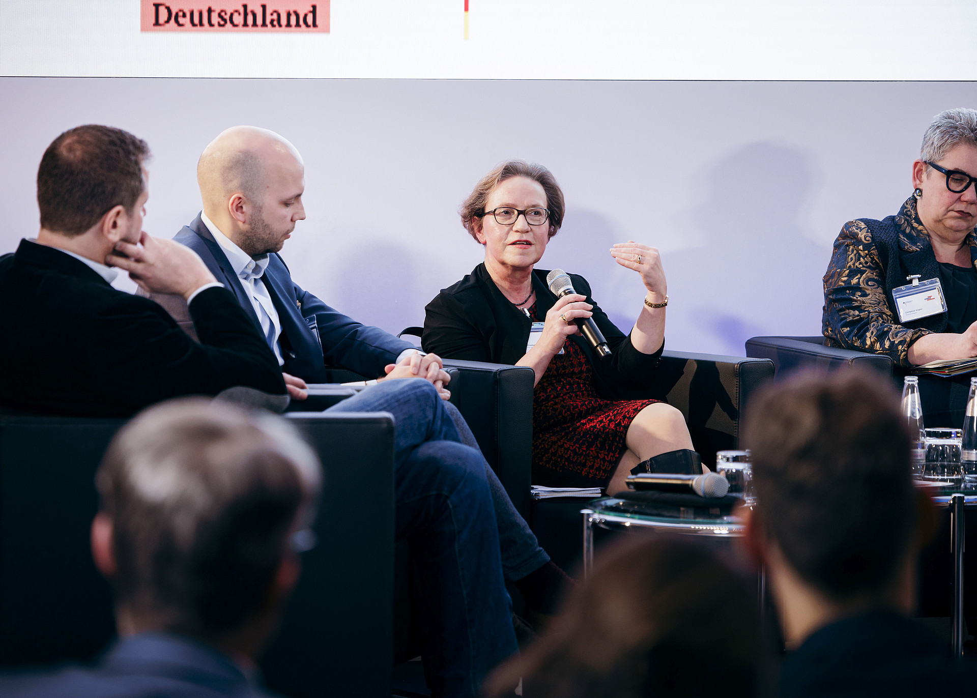 Vier Personen sitzen bei einer Podiumsdiskussion. Eine Frau in der Mitte hält ein Mikrofon und gestikuliert beim Sprechen. Die anderen drei hören aufmerksam zu. Im Vordergrund ist ein Publikum zu sehen, und darüber wird auf einem Bildschirm ein Teiltext angezeigt.