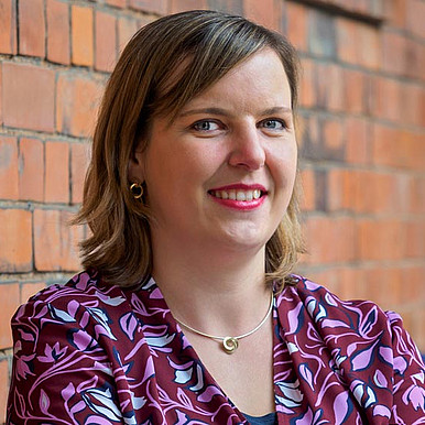 Eine Frau mit schulterlangem braunem Haar, einer gemusterten kastanienbraunen Bluse und Goldschmuck steht lächelnd mit verschränkten Armen vor einer Backsteinmauer.