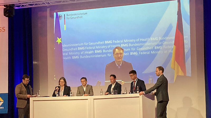 Fünf Personen sitzen an einem Podiumstisch mit Blick auf das Publikum. Hinter ihnen ist auf einem großen Bildschirm das Logo und der Name des Bundesministeriums für Gesundheit zu sehen. Auch die deutsche und die EU-Flagge sind zu sehen. Die Podiumsteilnehmer sind in ein Gespräch vertieft.