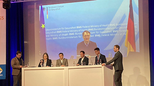 Fünf Personen sitzen an einem Podiumstisch mit Blick auf das Publikum. Hinter ihnen ist auf einem großen Bildschirm das Logo und der Name des Bundesministeriums für Gesundheit zu sehen. Auch die deutsche und die EU-Flagge sind zu sehen. Die Podiumsteilnehmer sind in ein Gespräch vertieft.
