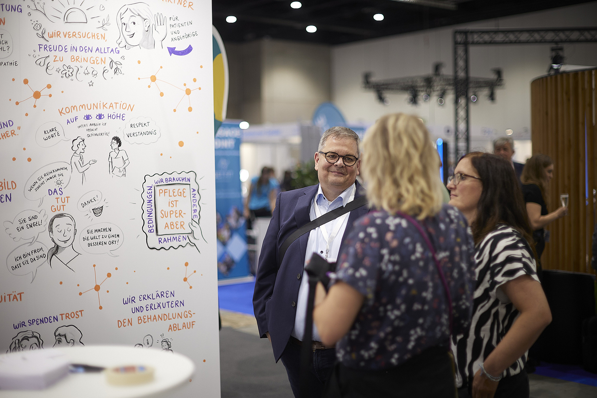 Ein Mann und zwei Frauen unterhalten sich auf einer Veranstaltung. Hinter ihnen hängt ein großes, illustriertes Plakat mit Texten und Zeichnungen. Die Kulisse scheint ein Konferenz- oder Ausstellungssaal zu sein.