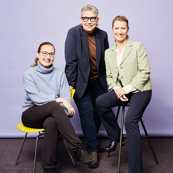 Drei Personen posieren lächelnd vor einem hellvioletten Hintergrund. Zwei sitzen auf gelben Hockern, eine steht in der Mitte. Alle tragen Brillen und Business-Casual-Kleidung in Blau-, Grün- und Brauntönen.