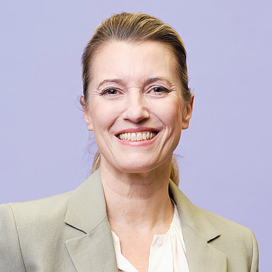 Eine lächelnde Frau mit zurückgekämmtem hellem Haar, die einen hellgrauen Blazer und eine weiße Bluse trägt, steht vor einem schlichten hellvioletten Hintergrund.