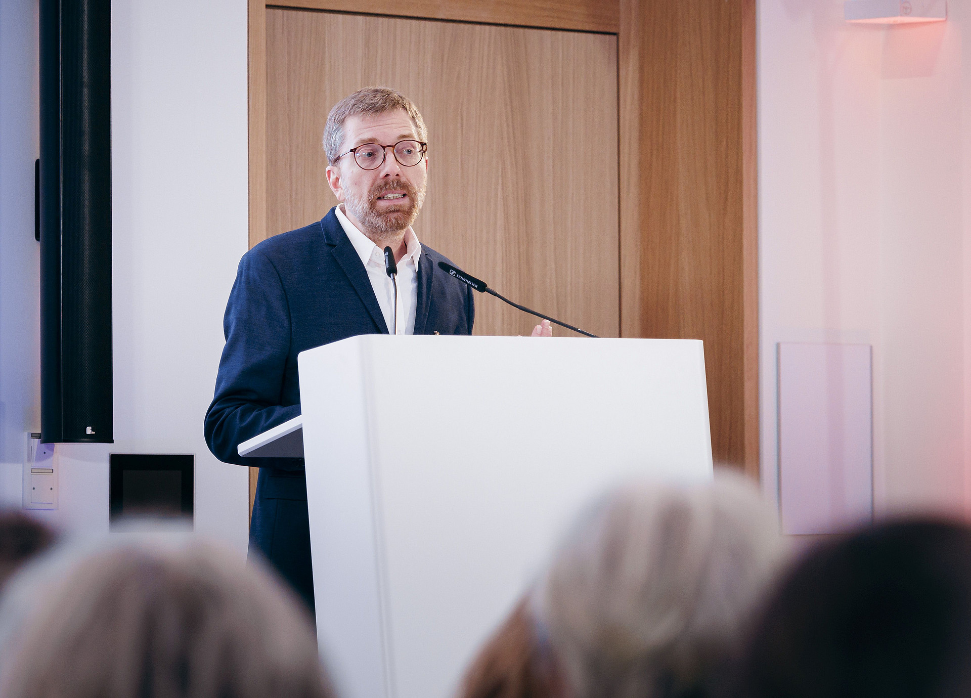 Ein Mann in Anzug und Brille spricht an einem Podium. Er befindet sich in einem gut beleuchteten Raum mit einer Holztür hinter ihm. Der Fokus liegt auf ihm, im Vordergrund ist ein Publikum teilweise sichtbar.