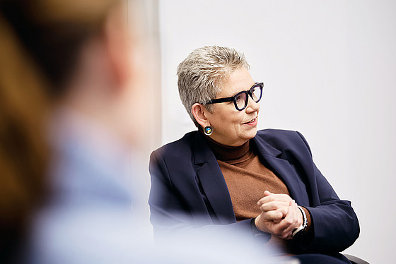 Eine ältere Person mit kurzen grauen Haaren, Brille und marineblauem Blazer sitzt mit gefalteten Händen da und hört aufmerksam zu. Der Hintergrund ist schlicht, eine weitere Person ist im Vordergrund unscharf zu sehen.