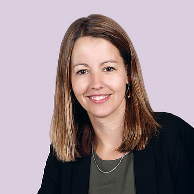 Eine Frau mit glattem, schulterlangem hellbraunem Haar lächelt in die Kamera. Sie trägt einen dunklen Blazer über einem grünen Oberteil, eine Halskette und Creolen vor einem hellvioletten Hintergrund.