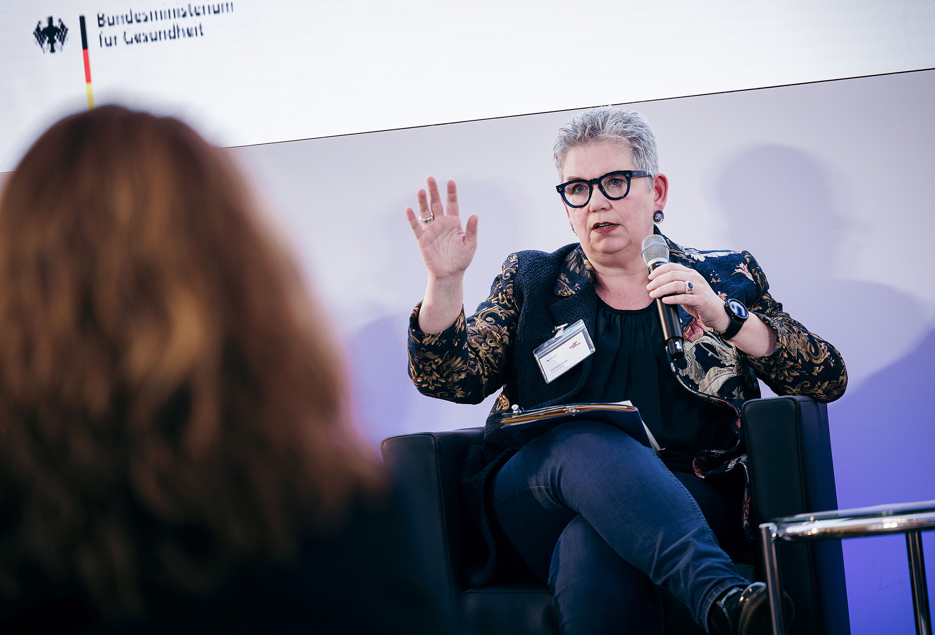 Eine Person mit kurzen grauen Haaren und Brille sitzt auf einem Stuhl, hält ein Mikrofon und gestikuliert mit einer Hand. Es ist Christine Vogler. ie trägt einen gemusterten Blazer und dunkle Kleidung. Eine weitere Person mit langen Haaren ist im Vordergrund zu sehen, leicht verschwommen.