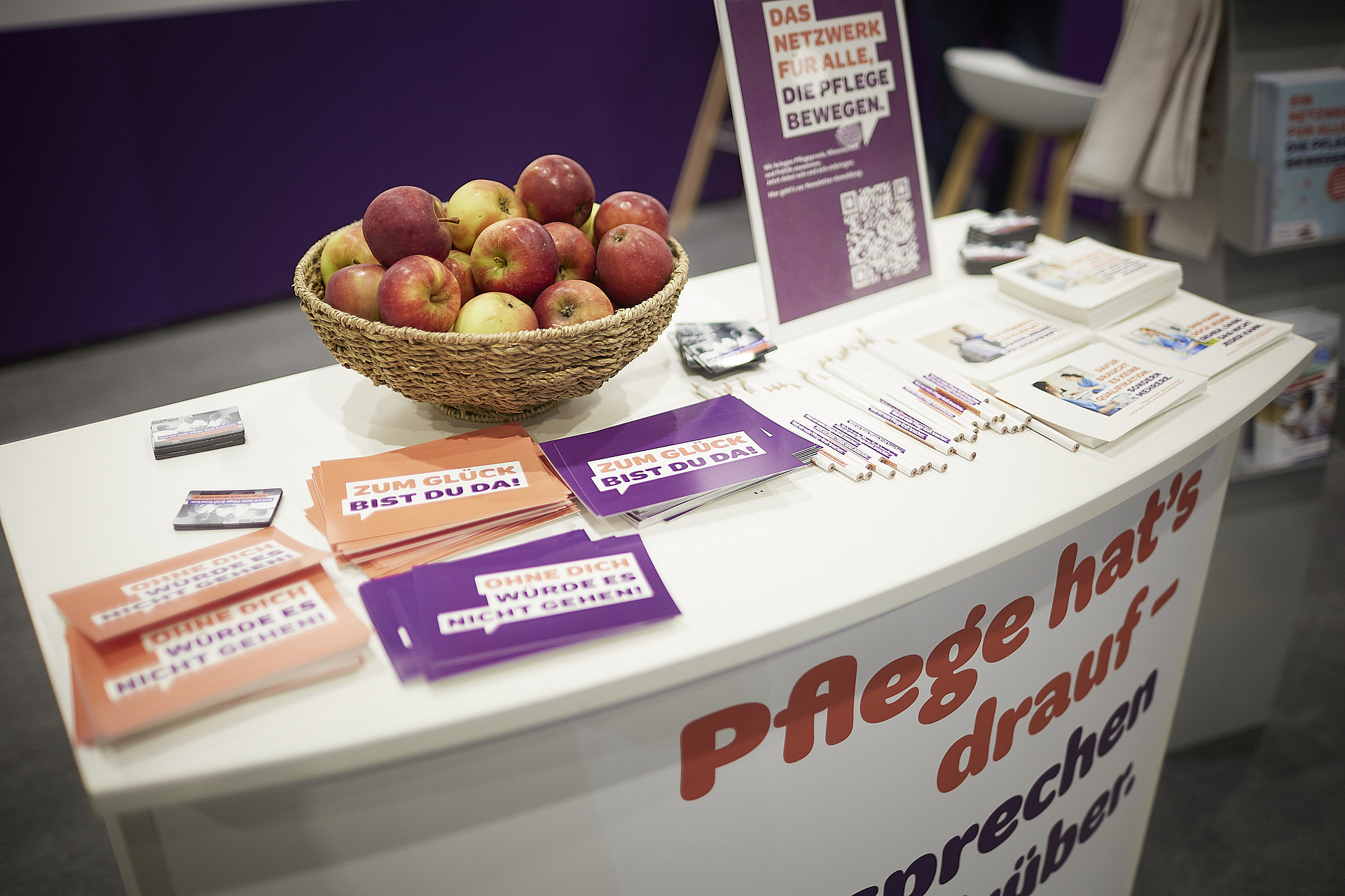 Ein Standtisch mit einem Korb Äpfel und verschiedenen Flyern auf Deutsch. Auf der Tischdecke ist teilweise Text zu sehen, darauf liegen Stifte und Broschüren. Hinter dem Tisch steht ein Schild mit weiteren Informationen.