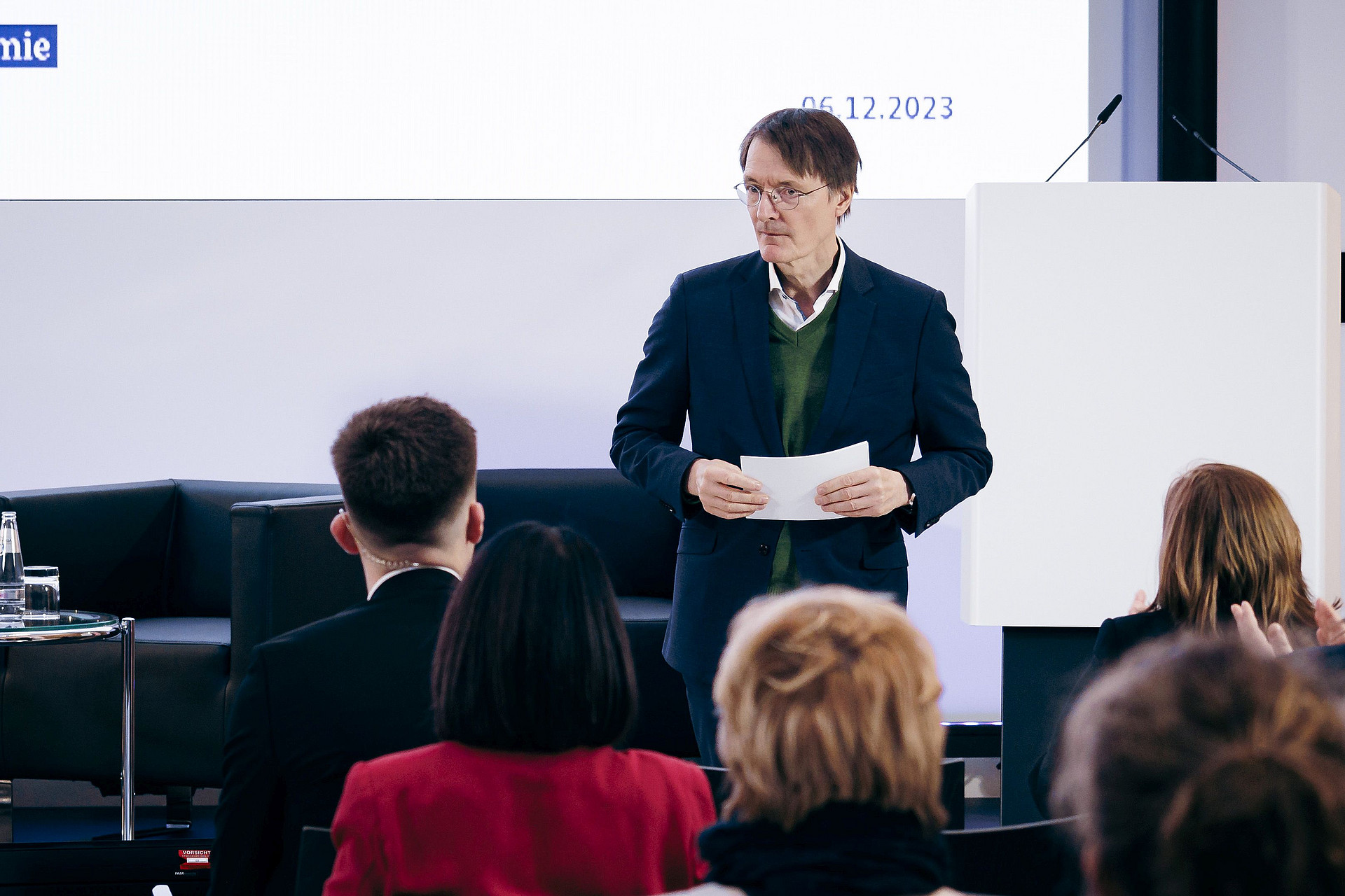 Eine Person in formeller Kleidung steht vor einem Publikum und hält ein Papier in der Hand. Es ist Prof. Karl Lauterbach. Sie steht neben einem weißen Rednerpult und auf einem großen Bildschirm wird das Datum 06.12.2023 angezeigt. Das Publikum scheint zu sitzen und aufmerksam zu sein.