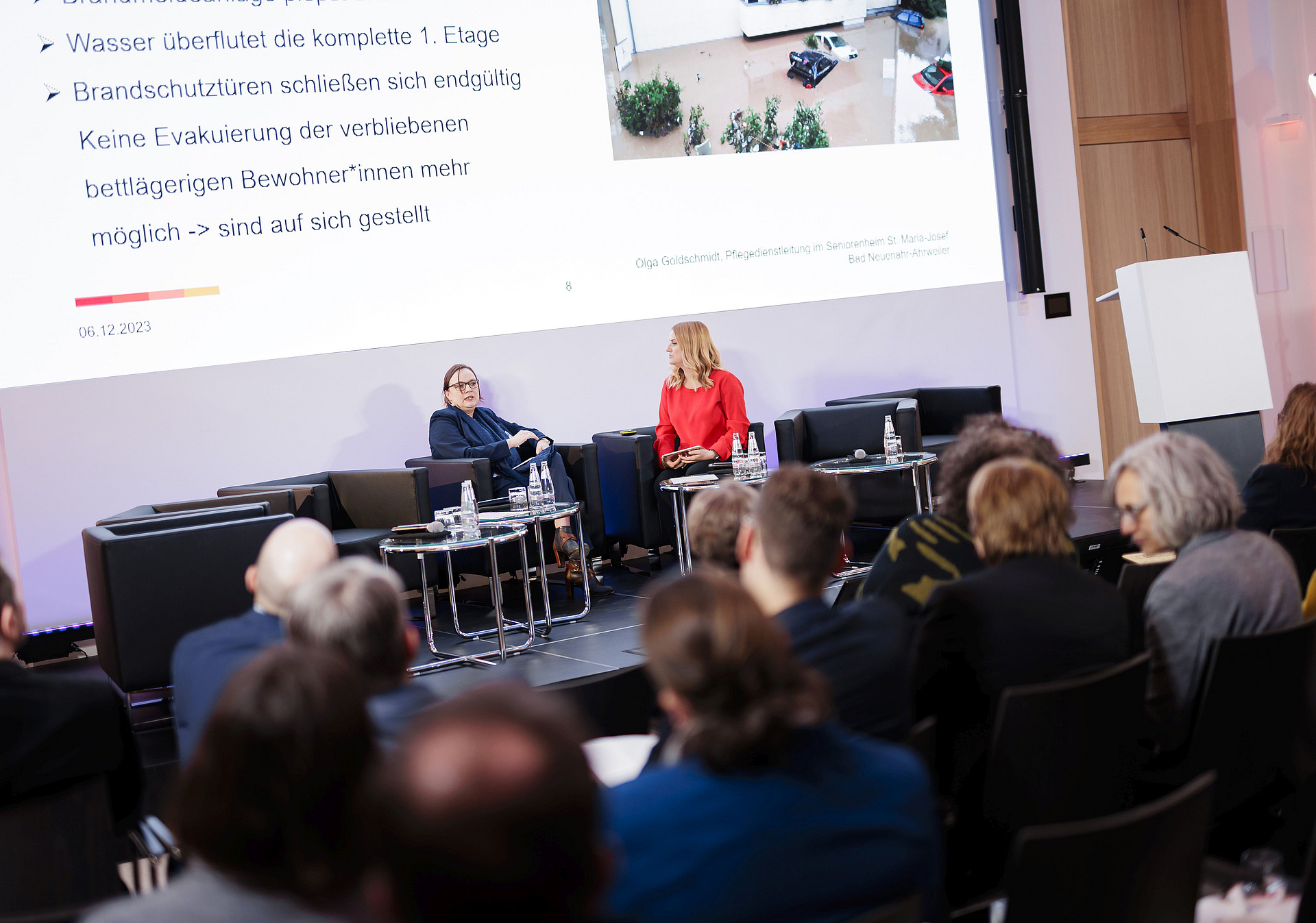 Ein Redner im roten Outfit und ein weiterer im dunklen Anzug sitzen auf der Bühne einer Konferenz. Ein Publikum beobachtet sie. Hinter ihnen ist ein großer Bildschirm, auf dem deutscher Text angezeigt wird.