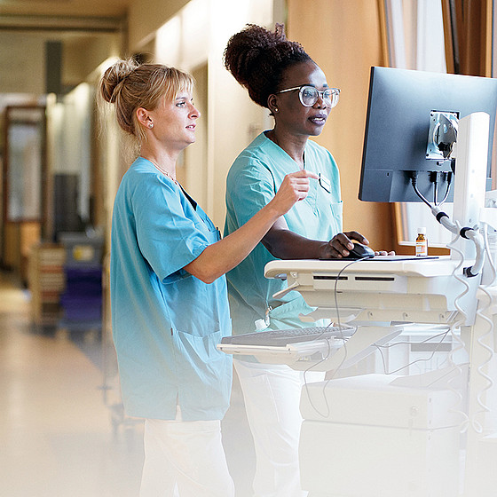 Zwei medizinische Fachkräfte in OP-Kleidung stehen an einem Computerarbeitsplatz in einem Krankenhausflur, überprüfen Informationen auf dem Monitor und besprechen etwas miteinander.