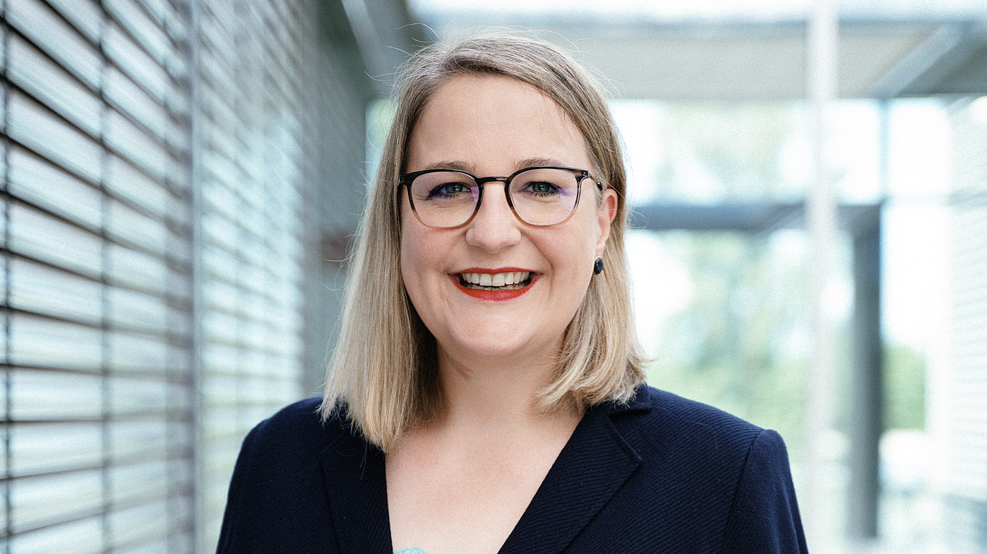 Eine lächelnde Frau mit Brille und dunklem Blazer steht in einem modernen Gebäude mit Glas- und Metallakzenten. Es handelt sich um Frau Prof. Hiestand. Sie hat schulterlanges blondes Haar und trägt ein hellblaues Oberteil. Im Hintergrund sind verschwommene Fenster und Strukturelemente zu sehen.