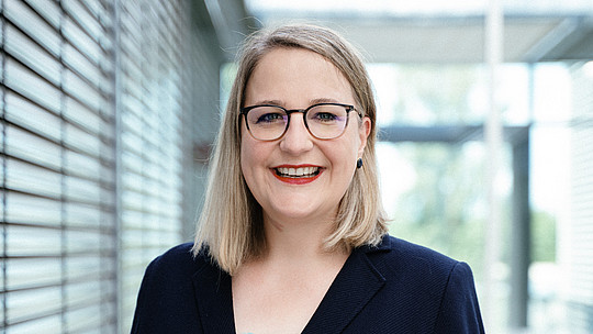 Eine lächelnde Frau mit Brille und dunklem Blazer steht in einem modernen Gebäude mit Glas- und Metallakzenten. Es handelt sich um Frau Prof. Hiestand. Sie hat schulterlanges blondes Haar und trägt ein hellblaues Oberteil. Im Hintergrund sind verschwommene Fenster und Strukturelemente zu sehen.