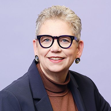 Lächelnde ältere Frau mit kurzen grauen Haaren, schwarzer Brille, braunem Rollkragenpullover, dunklem Blazer und runden Ohrringen, posiert vor einem hellvioletten Hintergrund.