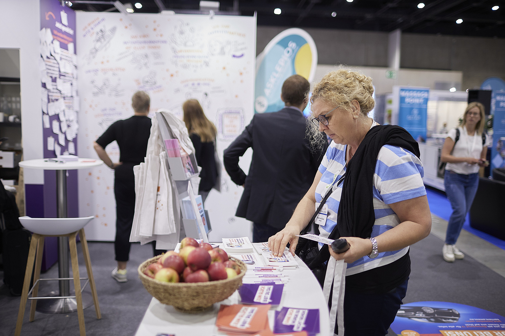 Eine Frau ordnet Materialien an einem Messestand. Auf dem Tisch steht ein Korb mit Äpfeln und Werbeartikeln. Im Hintergrund erkunden Besucher unter hellem Licht andere Stände und Ausstellungen.